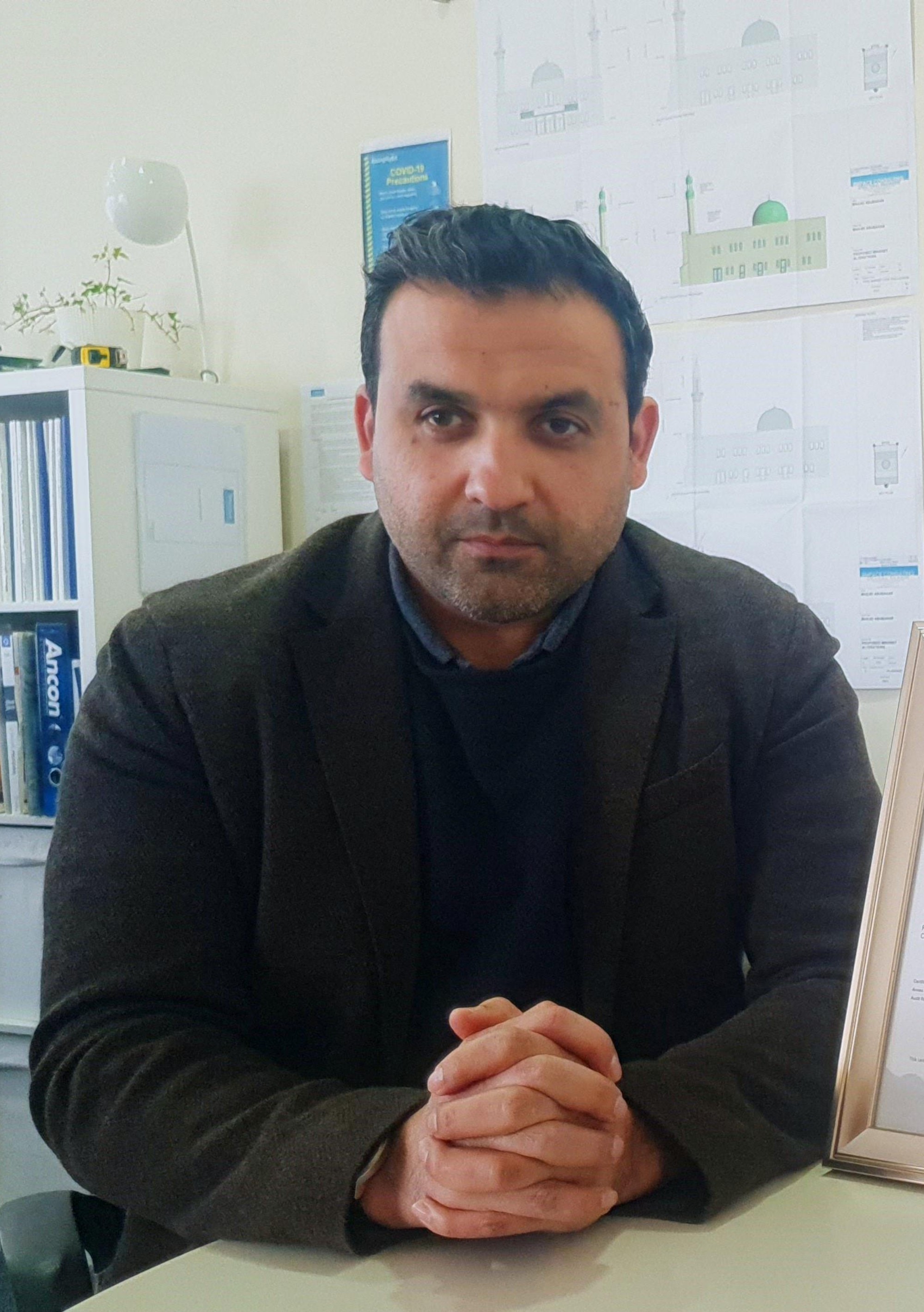 Portrait photo of Ummer sitting at a desk with his hands clasped, he is wearing smart workwear in a dark colour