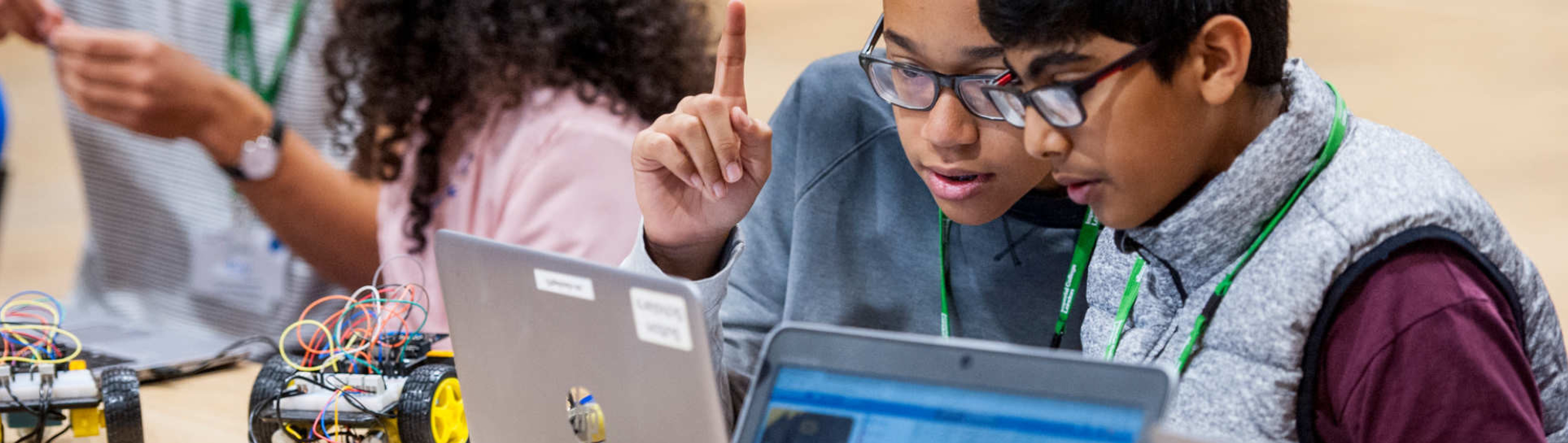 Two young students look at a laptop screen with a robot with wheels beside them.