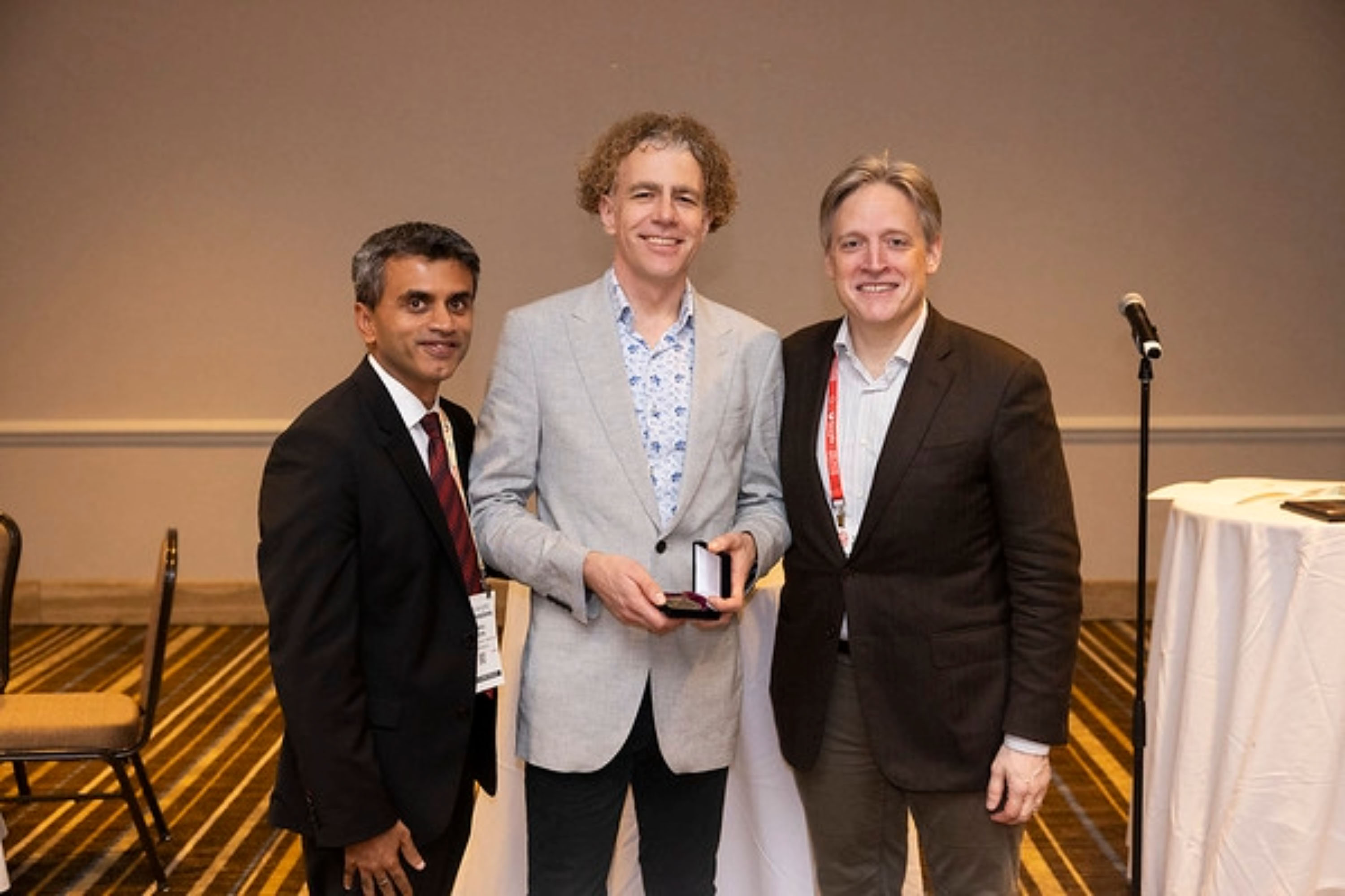 Three men in stand together smiling at the camera. The man in the centre is Professor James Ware, who has won an AHA Award, which he is holding.