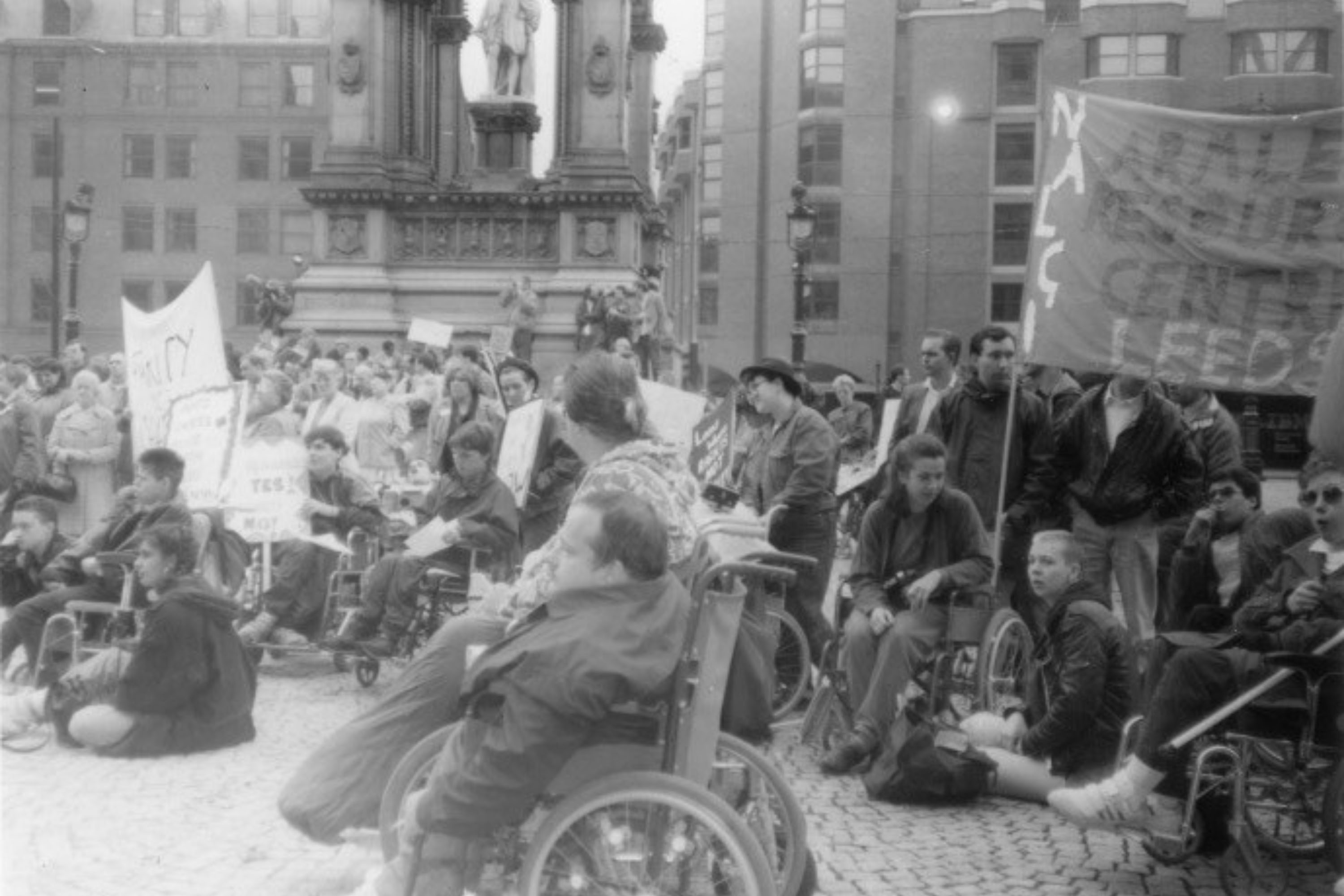 A view of a large crowd of people in Albert Square, Manchester; around the Albert memorial statue. Most of the crowd are looking to the left of the photo. On the far right is a banner which says ‘Armley Resource Centre Leeds’, only the letters ‘Armle Resour Centr Leed’ are visible. There is a mix of people standing and sitting on the ground or in wheelchairs. some are holding banners and placards.